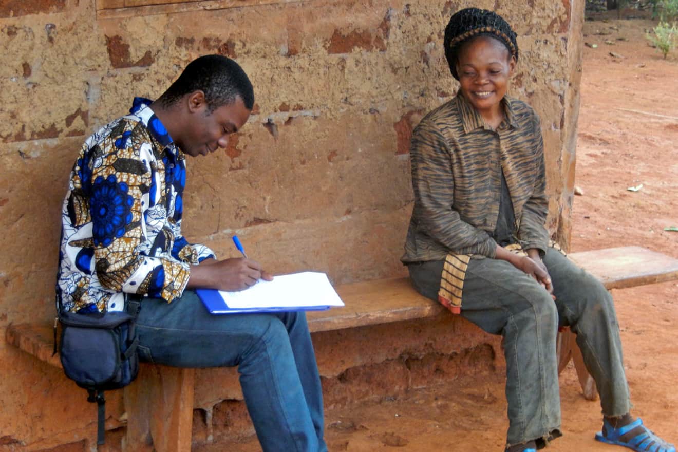 Ein Mann mit einem Schreibblock und eine Frau sitzen lächelnd auf einer Holzbank vor einer grob verputzten Wand