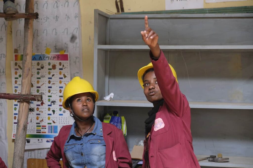 Eine angehende Solartechnikerin im roten Kittel und mit gelbem Helm zeigt ihrer Kollegin etwas.