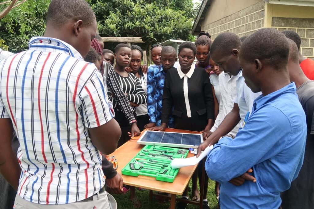 Die Teilnehmenden der Ausbildung sind um einen kleinen Tisch versammelt, auf dem ein Solarpanel und Werkzeug liegen.