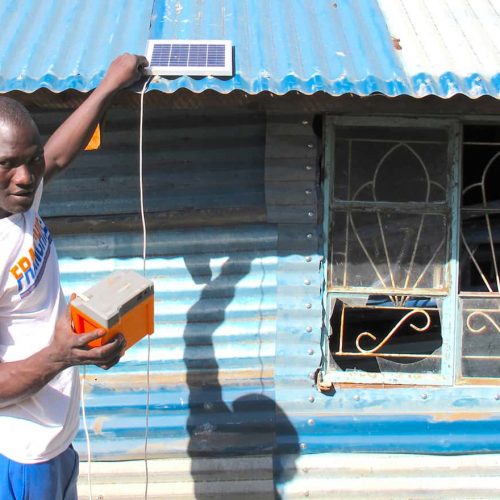 Ein junger Mann installiert ein kleines Solarpanel auf dem Dach eines blauen Wellblechhauses